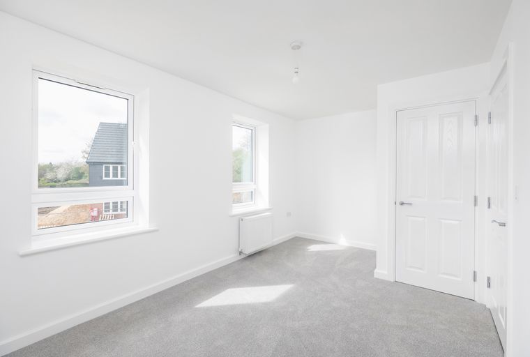 A bright, empty room with two windows, white doors, and a grey carpet.