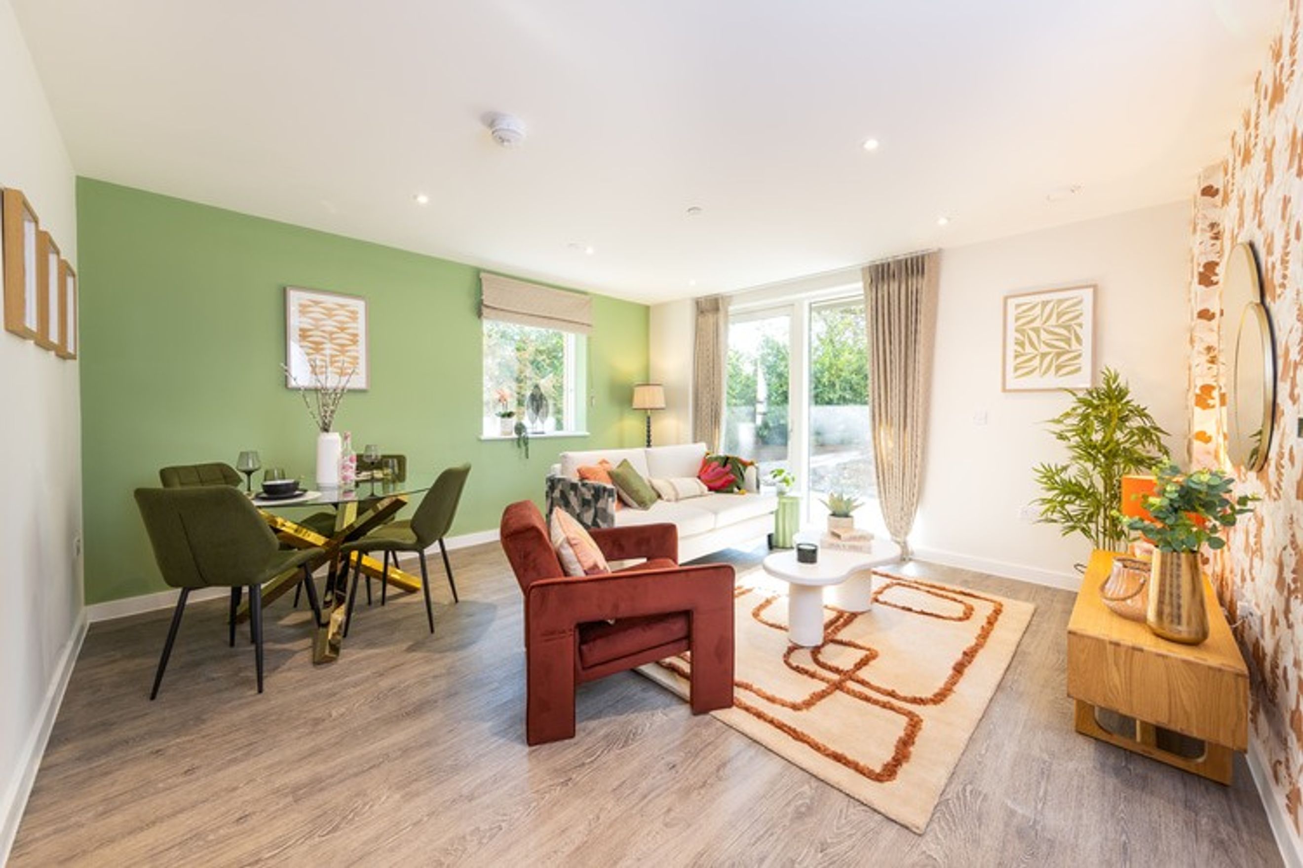 A dining and living area with a green wall, a glass dining table, a white sofa, and a rust-colored armchair.