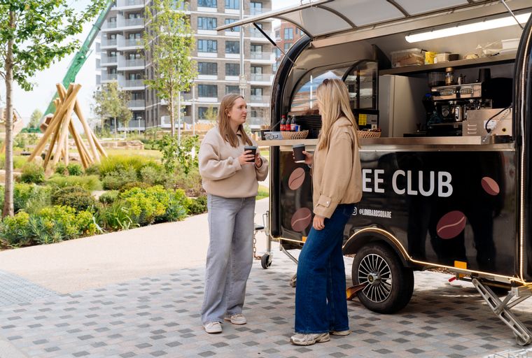 A coffee truck parked in front of two women, with modern apartment buildings in the background.