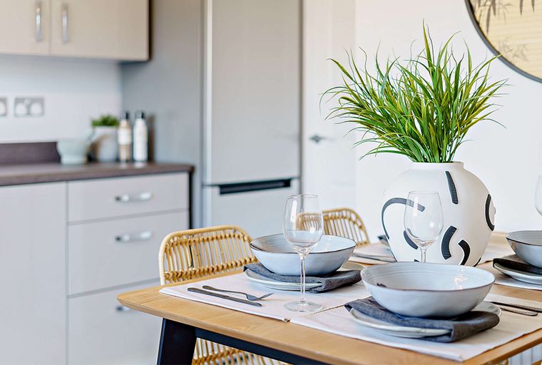 A dining table set for a meal with a vase of greenery and a kitchen in the background.