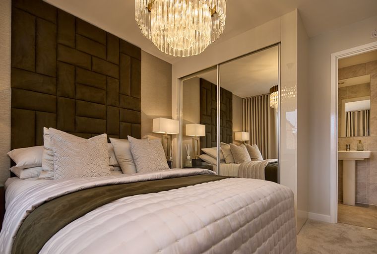A bedroom with a bed, headboard, lamps, chandelier, mirrored wardrobe, and an adjoining bathroom.