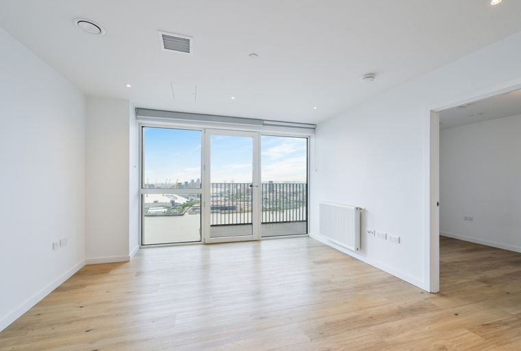 A modern, empty apartment room with large windows overlooking a city and water view, hardwood floors, and a white radiator.