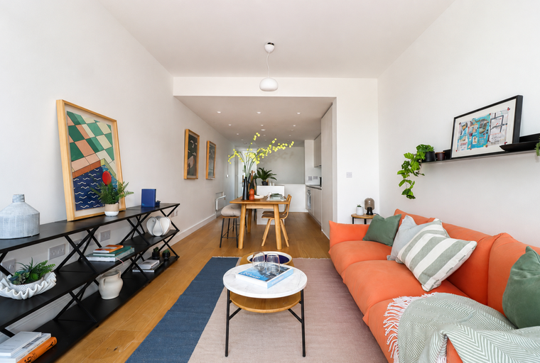 A living room and dining area with an orange sofa, wooden dining table, and a kitchen in the background.