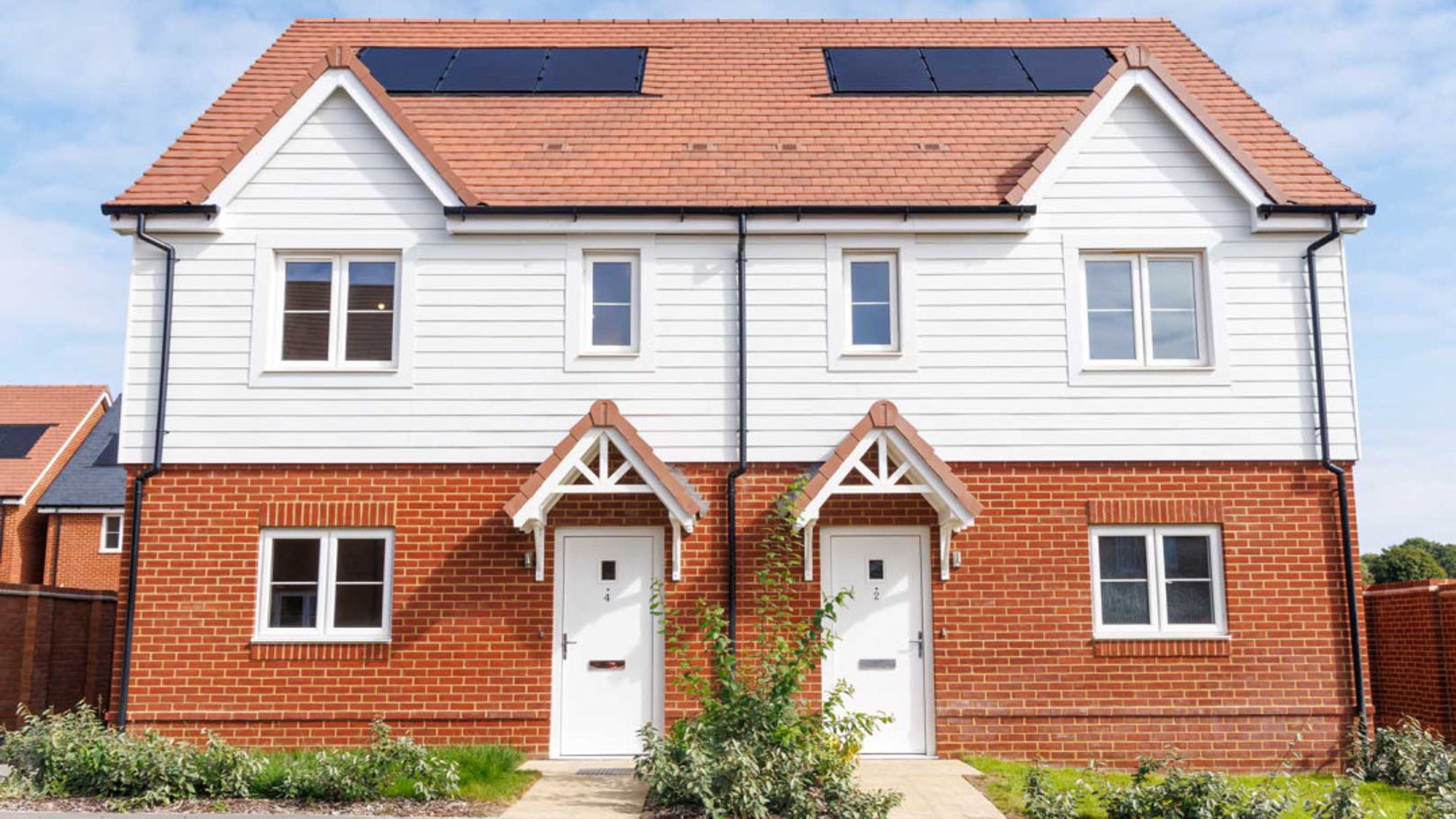 A pair of semi-detached houses with red brick lower floors and white siding upper floors, featuring white doors and windows, and solar panels on the roof.