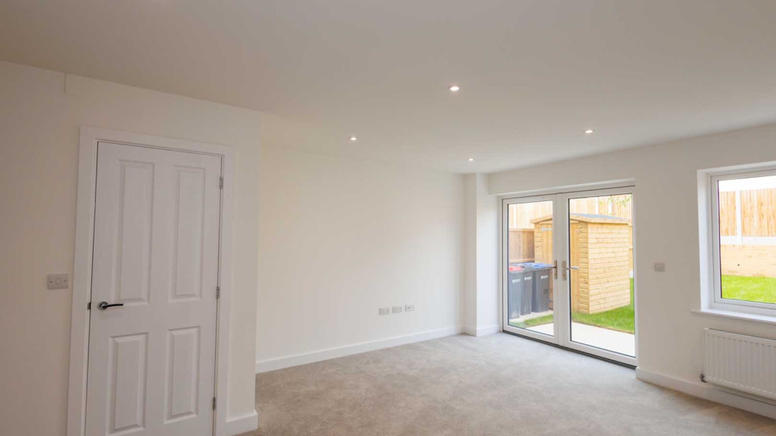 A room with a white door, grey carpet, and double glass doors leading outside.