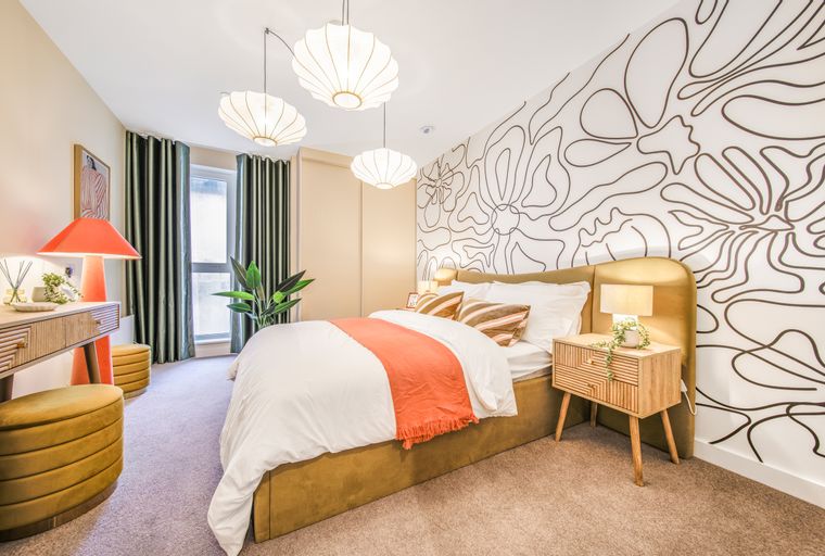 A bedroom with a bed, nightstand, vanity, and abstract wallpaper.