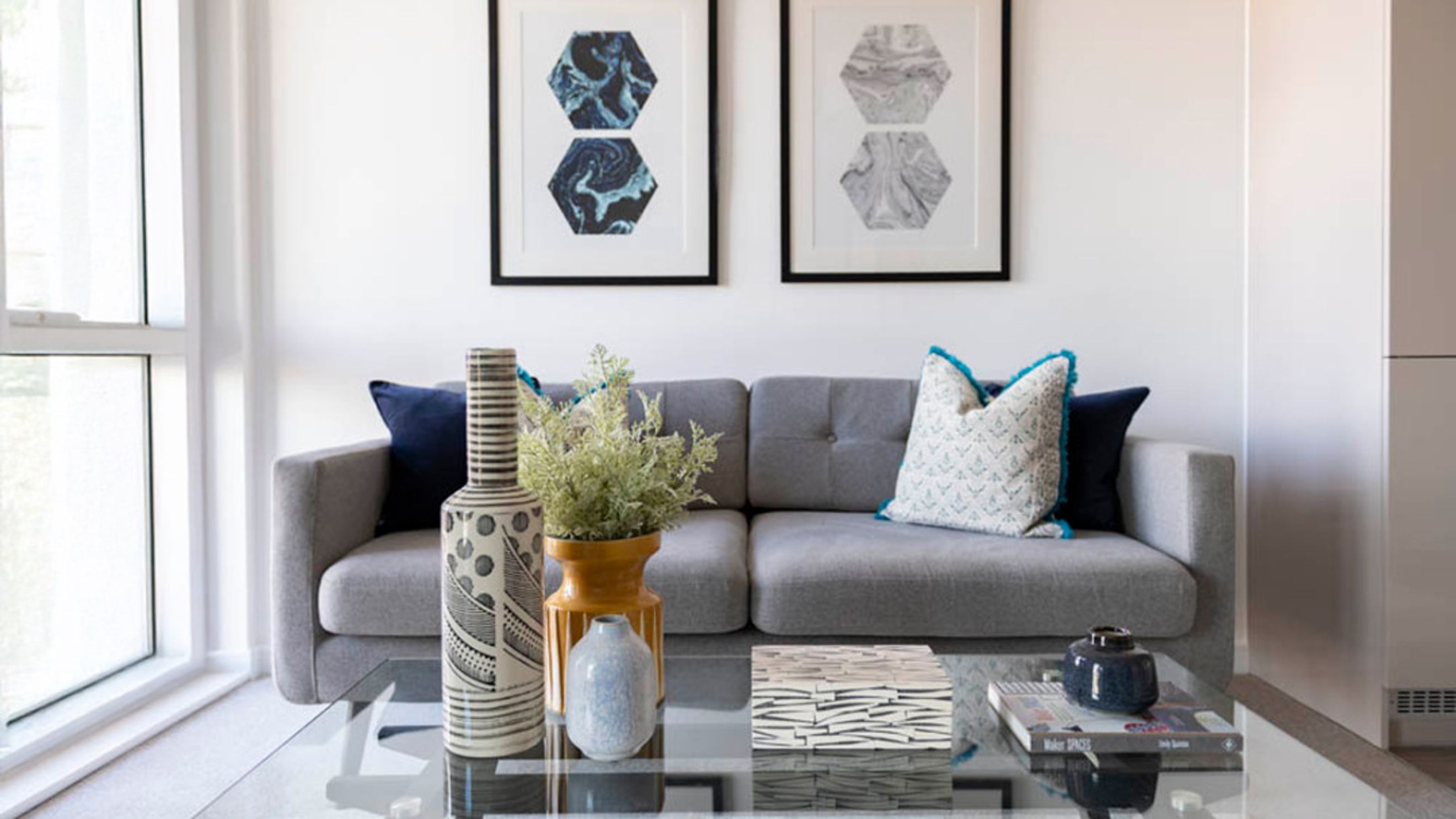 A living room with a grey sofa, decorative pillows, framed art, and a glass coffee table.