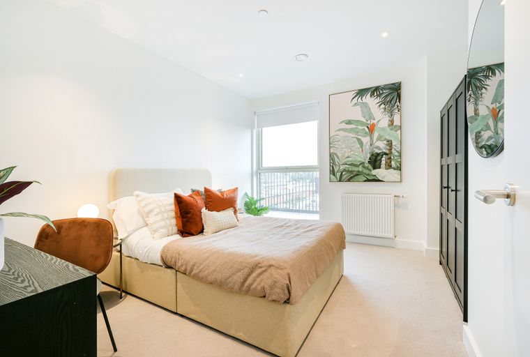 A bedroom with a bed, pillows, a throw, a chair, a desk, a window, artwork, and a wardrobe.