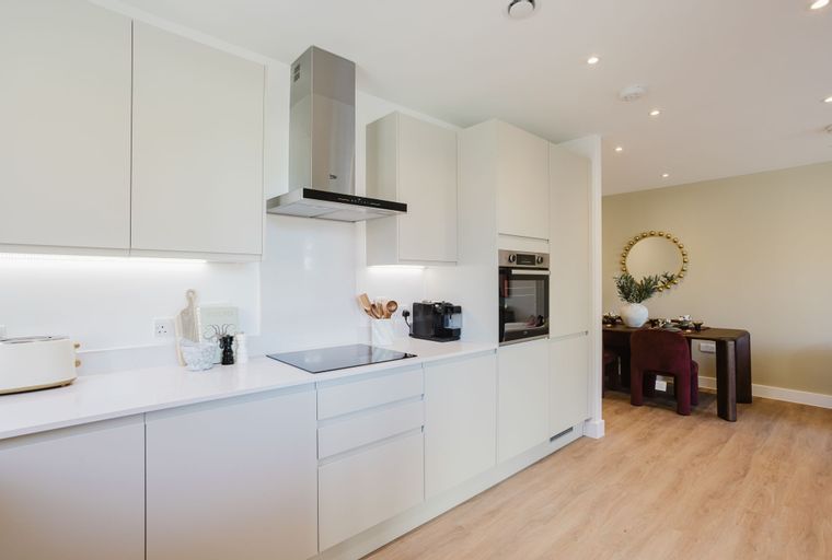 A modern kitchen with white cabinets, a stainless steel range hood, and a built-in oven, adjacent to a dining area with a table and chairs.