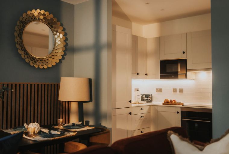 A dining table set for two with a lamp and a gold mirror above it, next to a kitchen with white cabinets and appliances.
