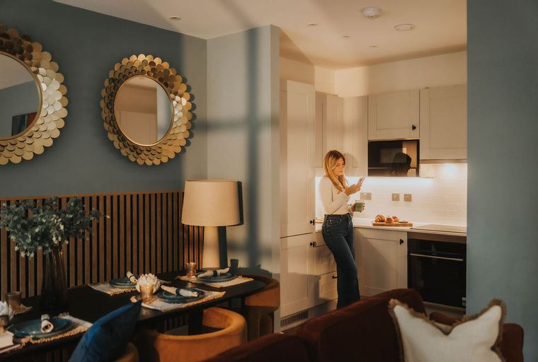 A woman stands in a modern kitchen, holding a phone and a cup, with a dining area visible in the foreground.