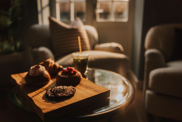 A wooden board with pastries and a drink on a glass table in front of armchairs.