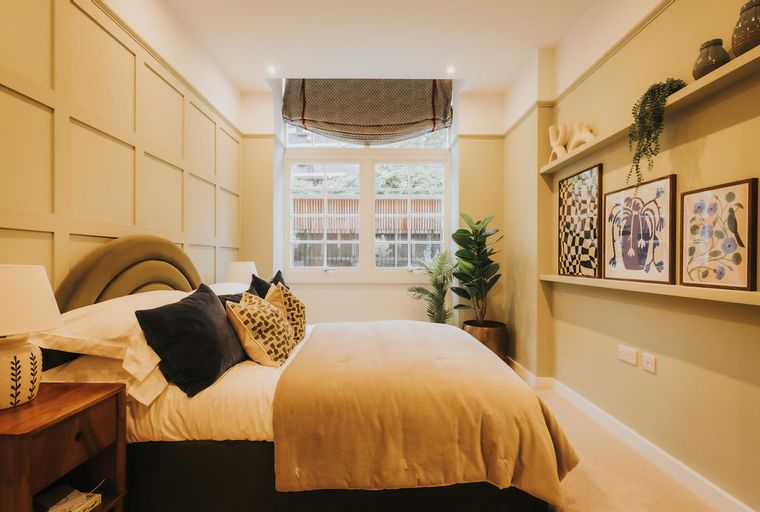A bedroom with a bed, pillows, lamps, a window, and artwork on shelves.