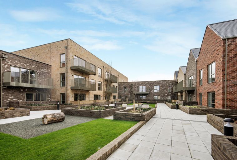 A courtyard with modern brick buildings, paved walkways, and landscaped garden beds.