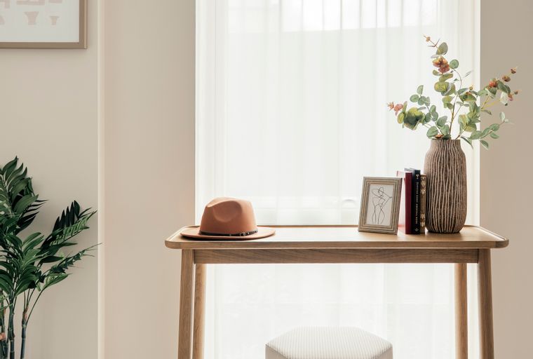 A wooden console table with a hat, books, a vase, and a framed picture sits in front of a window with sheer curtains, next to a plant and a striped stool.