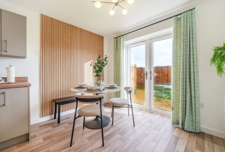 A dining area with a round table, chairs, and French doors leading to a garden, adjacent to a kitchen.