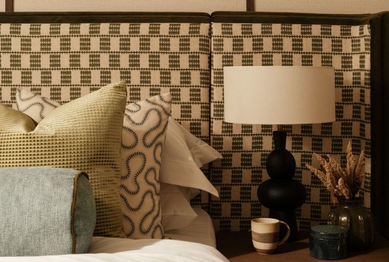 A bedroom with a patterned headboard, pillows, a lamp, and a bedside table.