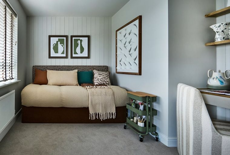 A bedroom with a daybed, pillows, artwork, a rolling cart, and a desk.