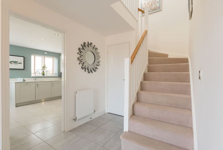 A hallway with a mirror, radiator, and a staircase leading to the upper level, with a view into a kitchen.