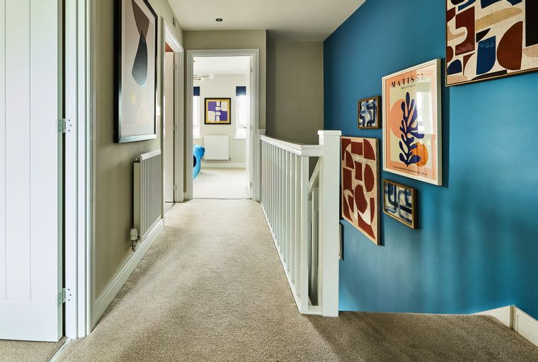 A hallway with a carpet, a blue wall with artwork, and a white banister.