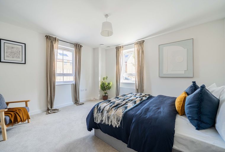 A bedroom with a bed, pillows, a throw blanket, windows, curtains, a chair, and artwork.