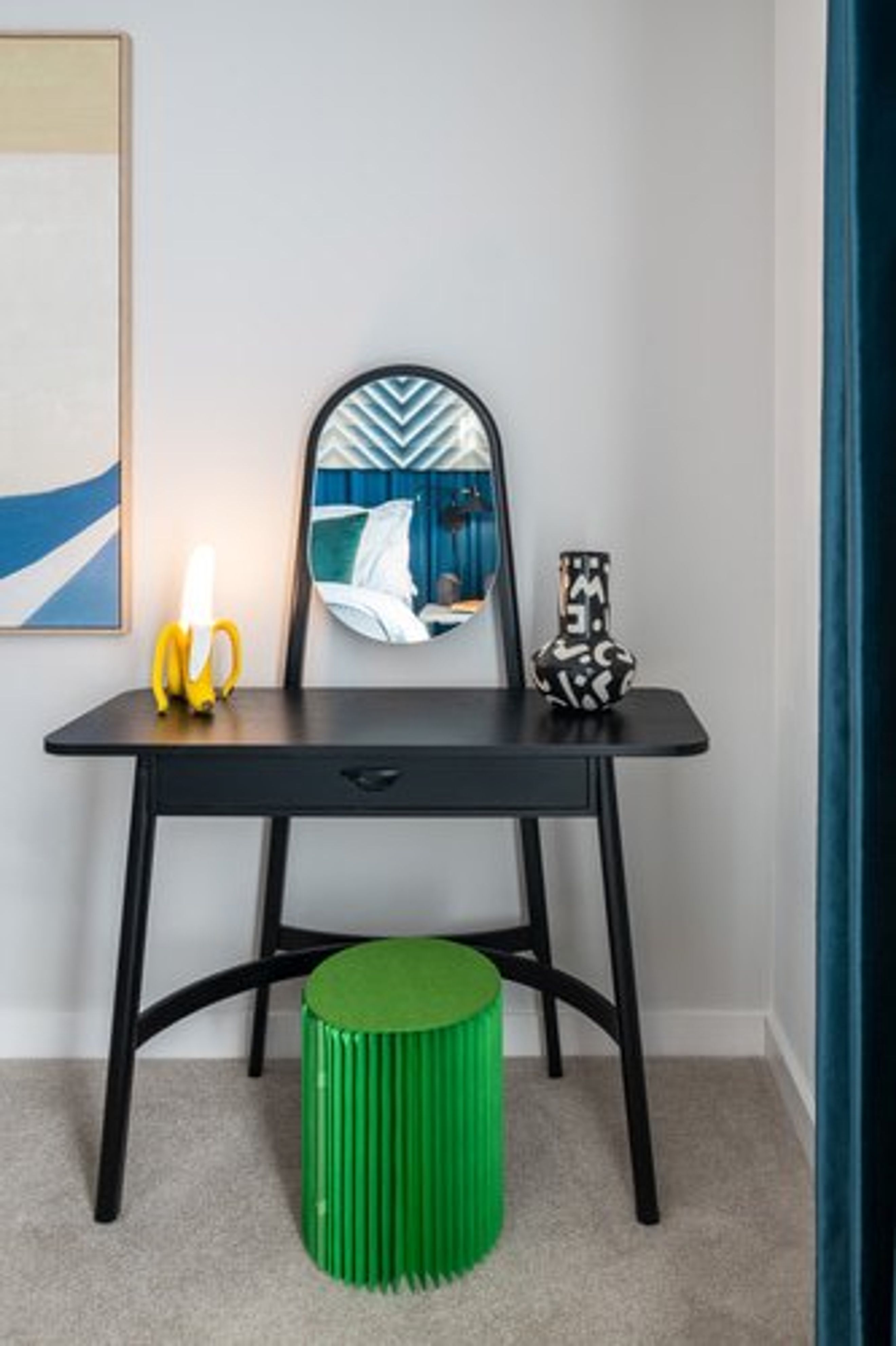 A black vanity desk with a mirror, a banana lamp, a vase, and a green stool.