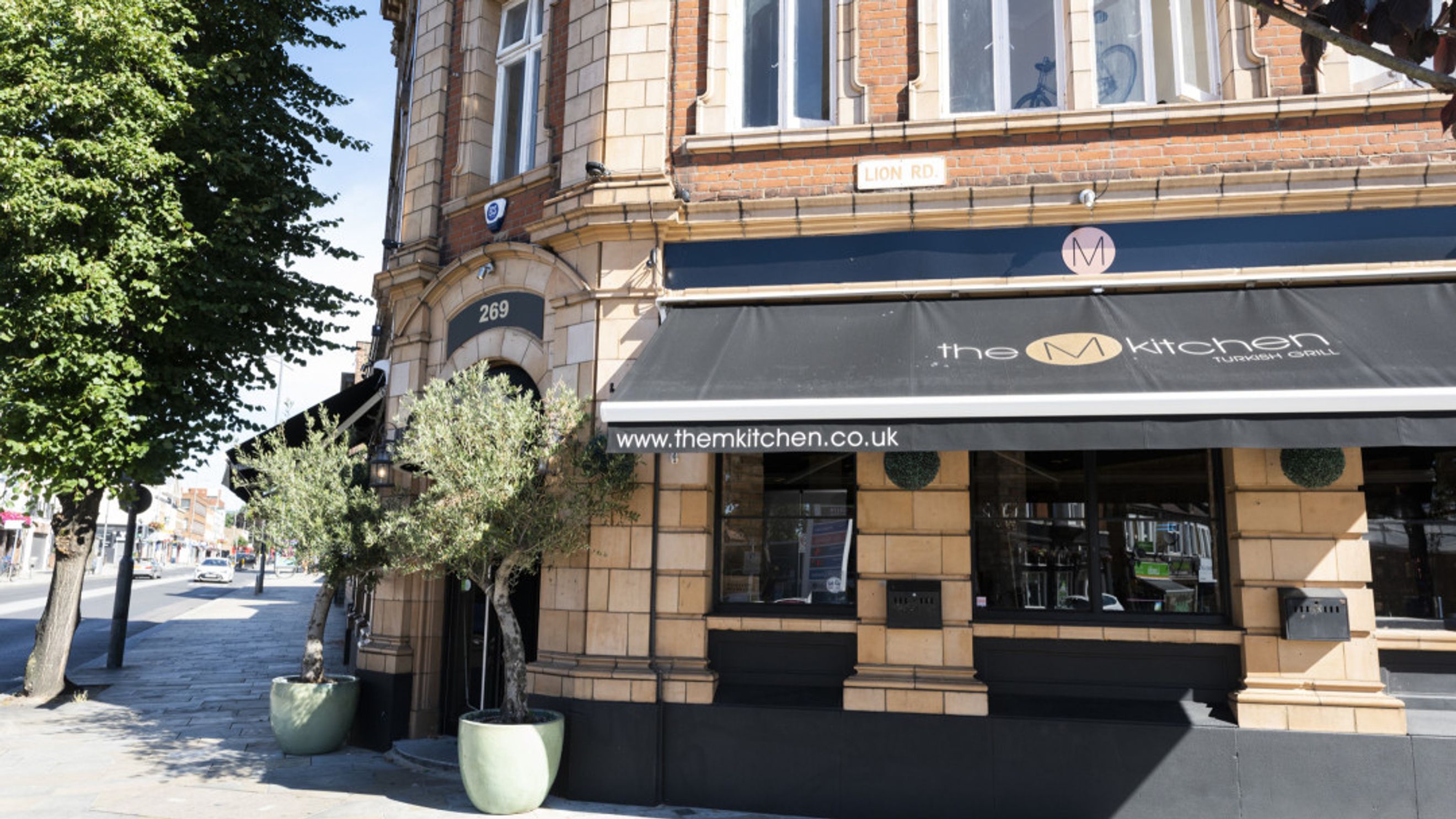 A storefront for 'The M Kitchen Turkish Grill' on a street corner with trees in pots.