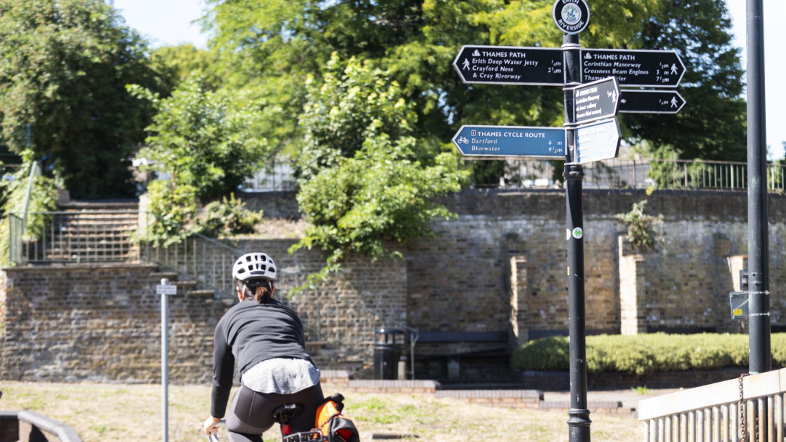 A cyclist wearing a helmet rides past a signpost indicating directions for the Thames Path and Thames Cycle Route.
