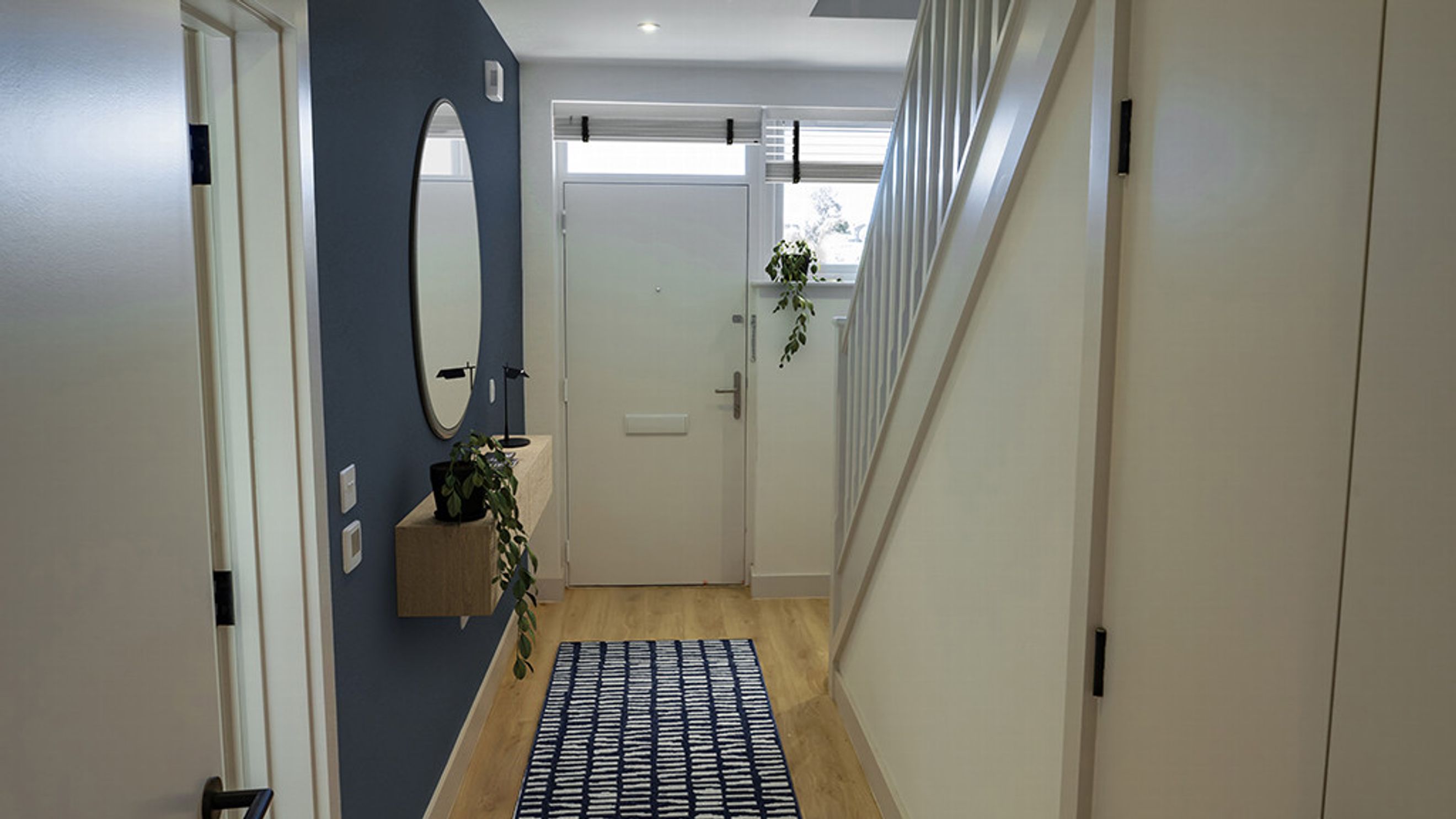 A hallway with a navy accent wall, an oval mirror, a floating shelf with a plant, a front door, and a staircase.