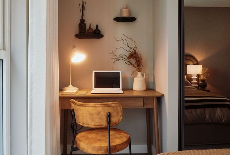 A desk with a laptop, lamp, and chair in front of decorative shelves, with a bedroom reflected in a mirror.