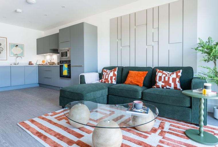 A living room and kitchen area with a green sectional sofa, a glass coffee table, and a grey kitchen.