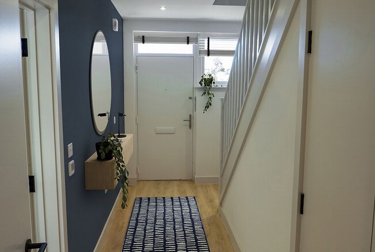 A hallway with a navy accent wall, an oval mirror, a floating shelf with a plant, a front door, and a staircase.