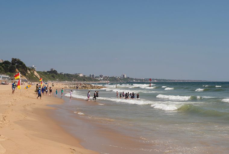 A sandy beach with people wading in the ocean and buildings on a distant cliffside.