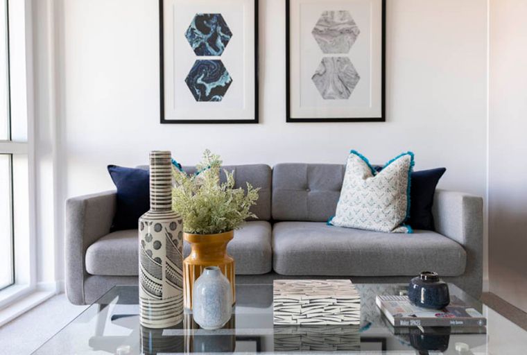 A living room with a grey sofa, decorative pillows, framed art, and a glass coffee table.