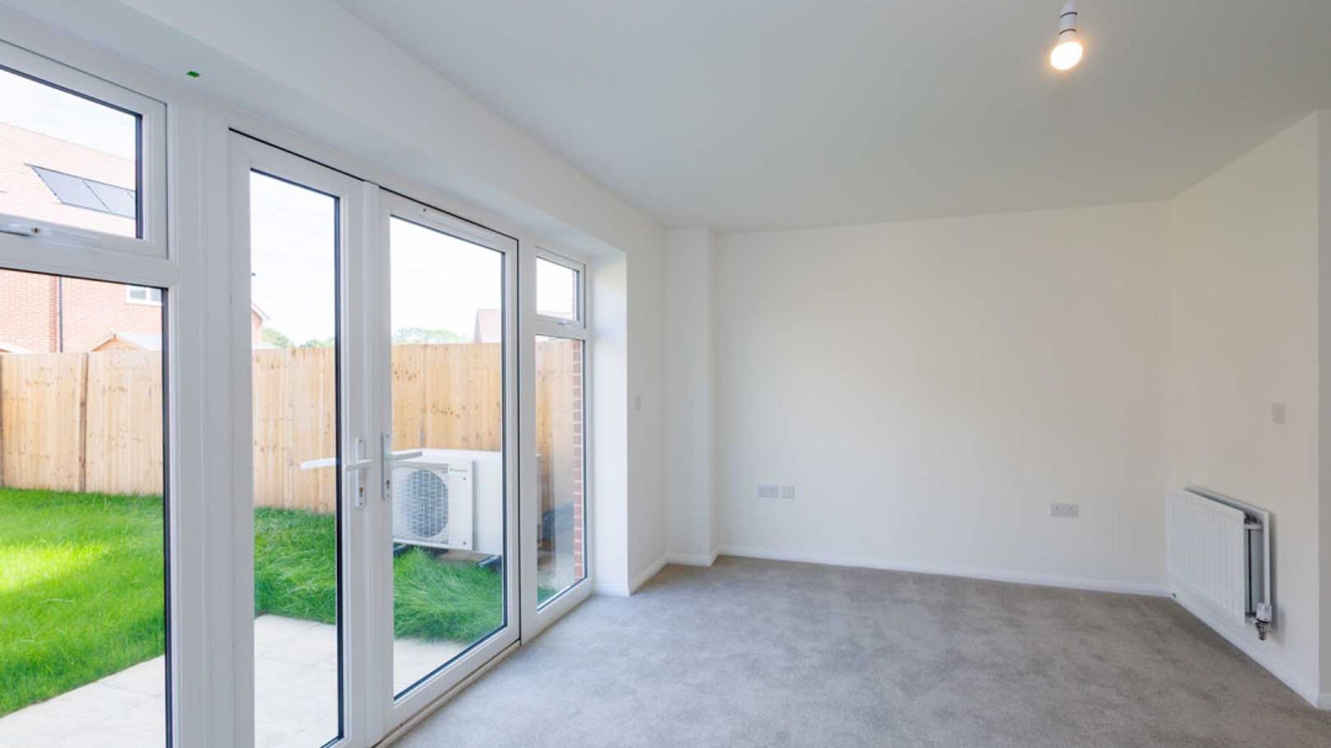 A living room with large glass doors opening to a garden, grey carpet, and a white radiator.