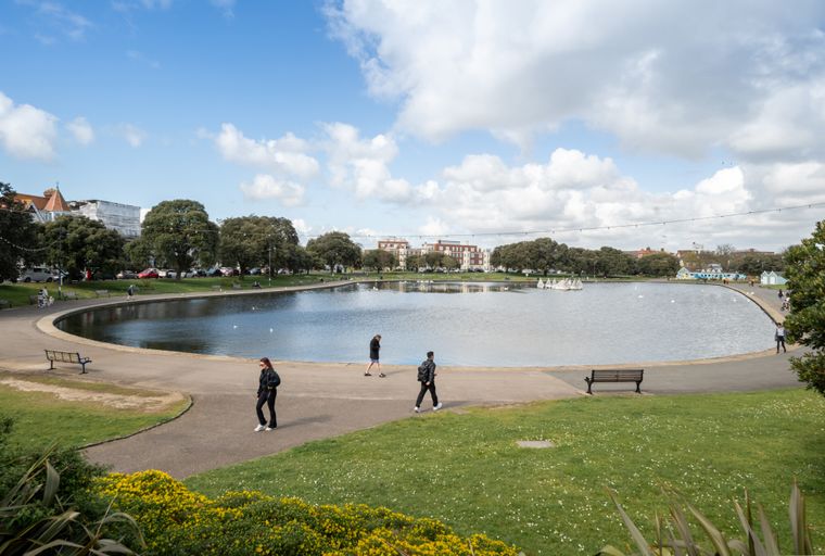 A park with a lake, trees, buildings, and people walking.
