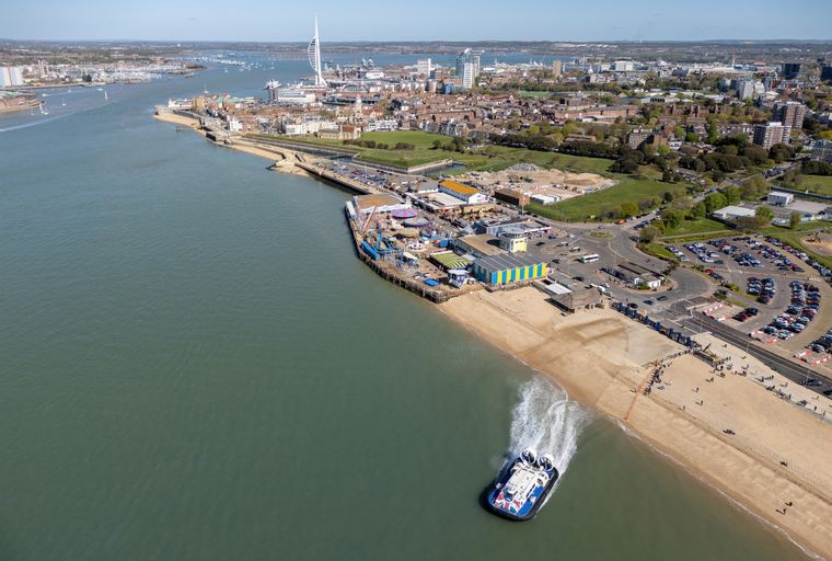 A hovercraft travels along the coast near a beach and a pier with amusement park rides.