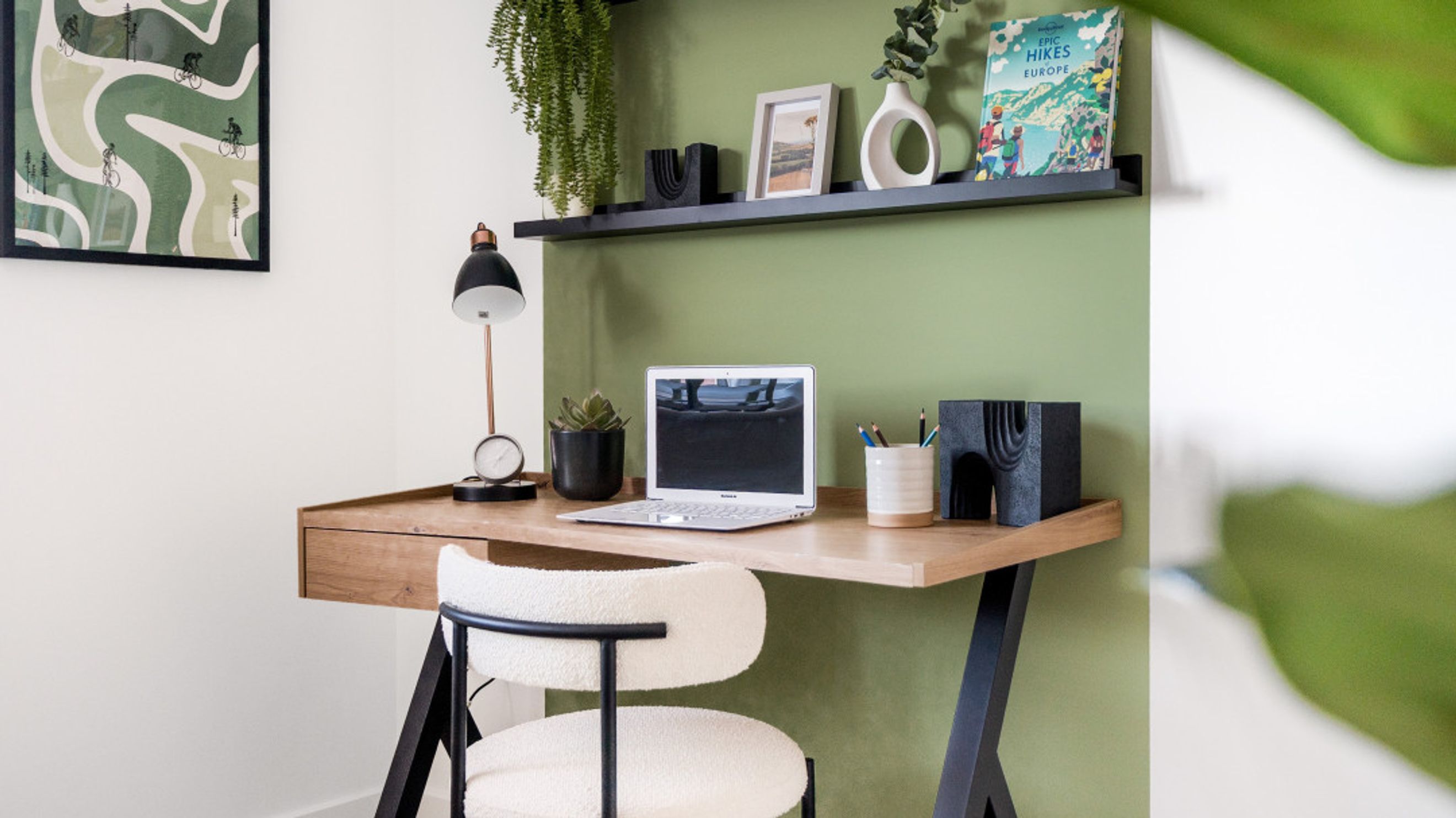 A home office desk with a laptop, chair, and decorative items on a green wall.