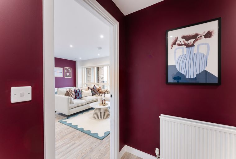 A hallway with a red wall and a framed picture, leading into a living room with a sofa and a coffee table.
