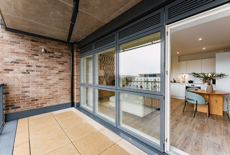 A balcony with a brick wall and large glass doors opening to a kitchen and dining area.